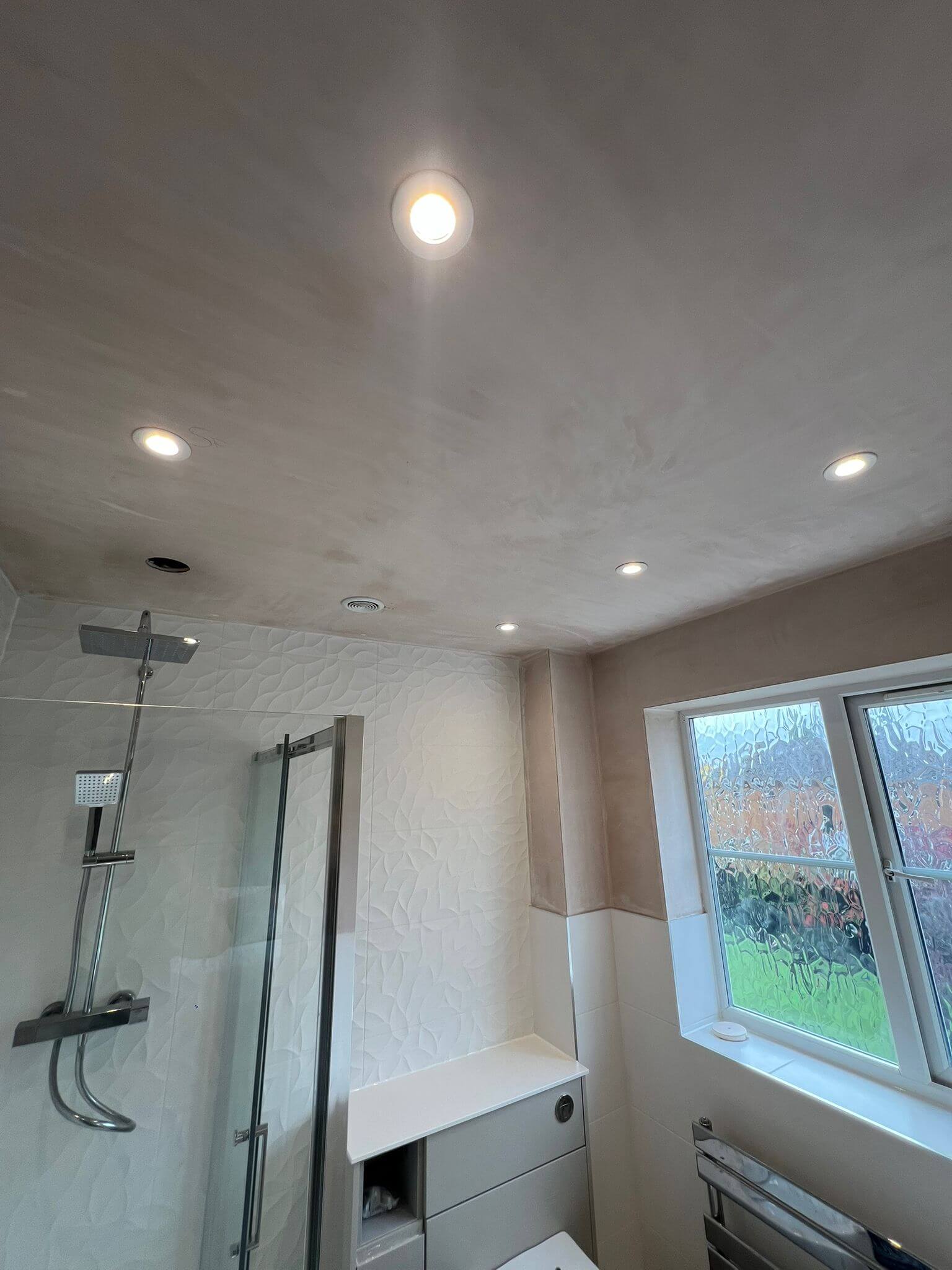 Interior view of a modern bathroom featuring chrome shower fixtures, textured white wall tiles, and a frosted window.